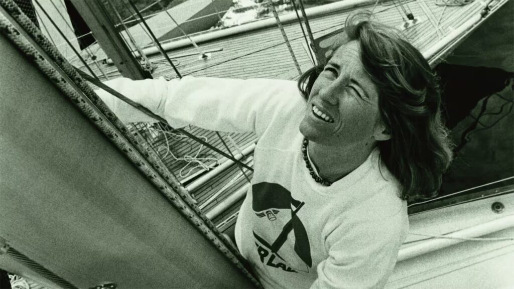 Naomi James in sailing gear on deck with rope lines and stormy sky in background.