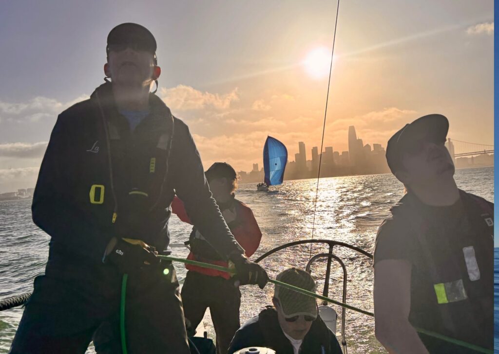 3 sailors aboard a yacht in the San Francisco bay at sunset, the city skyline visible behind